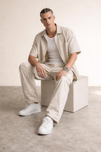 Man with braided hair sitting on white cube in beige shirt, pants, white tank top and sneakers