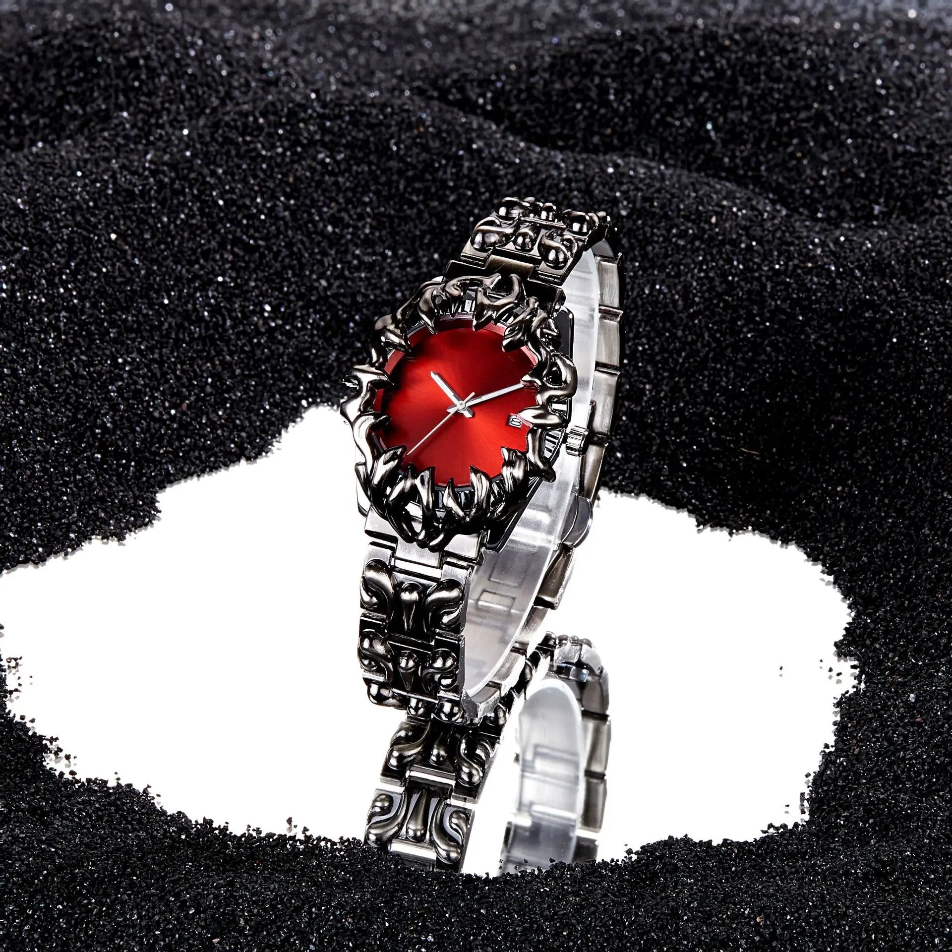 silver wristwatch with red dial and ornate flame-style bezel on reflective surface with black sand