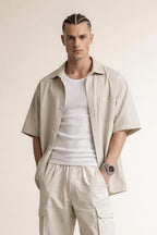 Man with braided hair wearing beige short-sleeve shirt, white tank top, beige cargo pants, and wristwatch