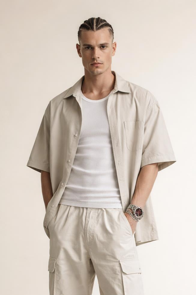 Man with braided hair wearing beige short-sleeve shirt, white tank top, beige cargo pants, and wristwatch